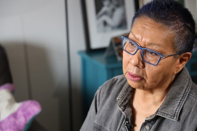 A woman with short hair and blue-rimmed glasses sits in a living room.