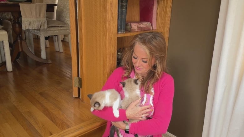 A woman in a bright pink shirt sits on the ground holding two kittens. 