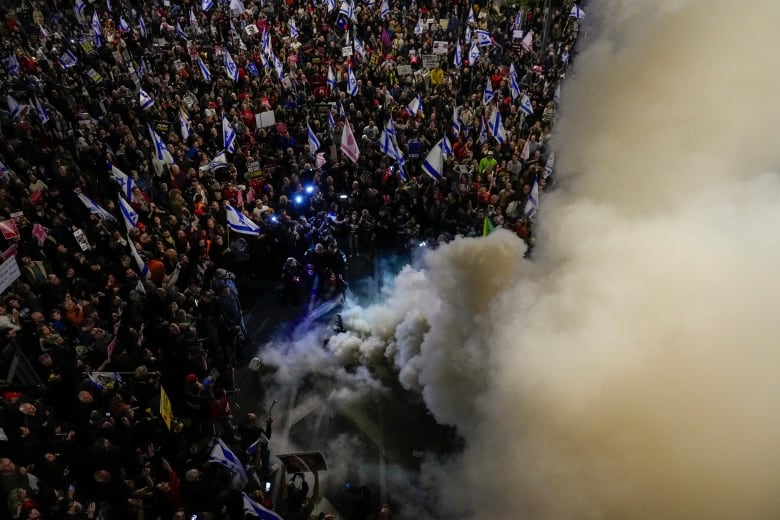 An aerial view shows protesters carrying flags and signs blocking a road while smoke rises.