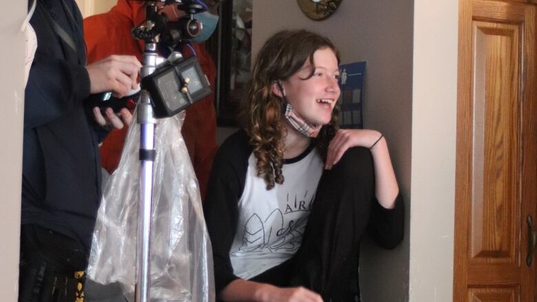 A teenage girls sits down and takes a break from directing. She is wearing a baseball shirt, with a mask under her chin, and holds a can of Pepsi.