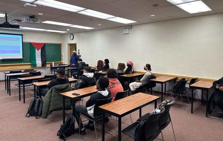 People in a classroom, with a Palestinian flag set up on a chalkboard.