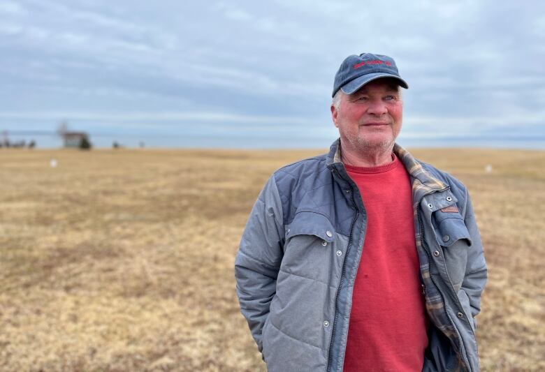 A man wearing a ballcap stands in an open field with the ocean in the background.