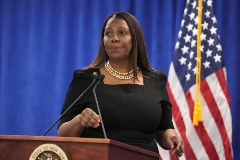 A dark complected woman wearing a dress and pearls is shown standing at a podium in front of an American flag.