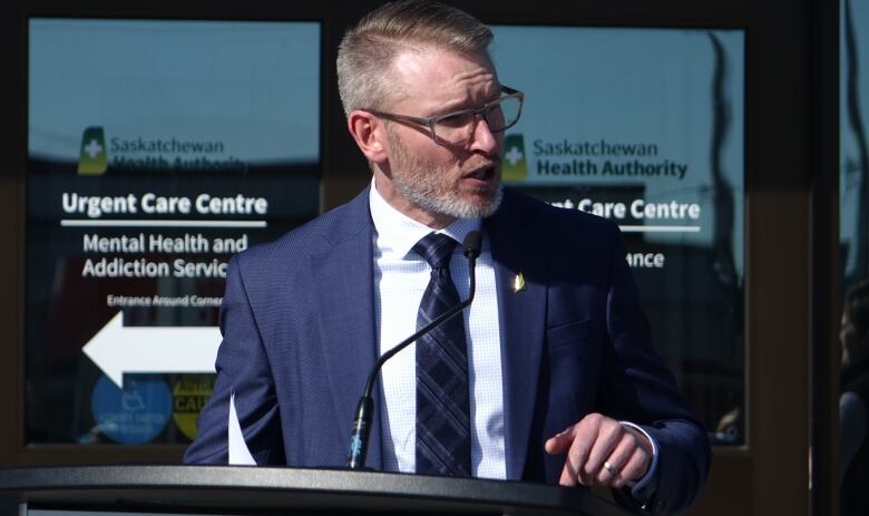 A man in a dark blue suit and glasses speaks at a podium.