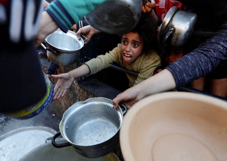 A child gestures as others in a crowd hold out empty pots at a food distribution site.