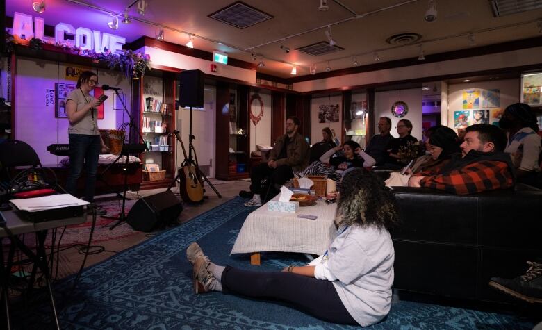 People sit on couches listening to a person standing at the front of the room and reading from a small notebook.