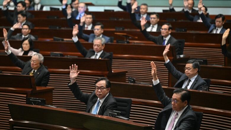 Several cleanshaven men in suits raise their arms while seated in what appears to be a legislative chamber.