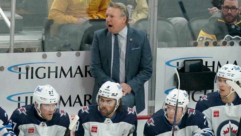 A hockey coach, wearing a suit, stands in the players box behind the players. He is shouting and looking to the left.