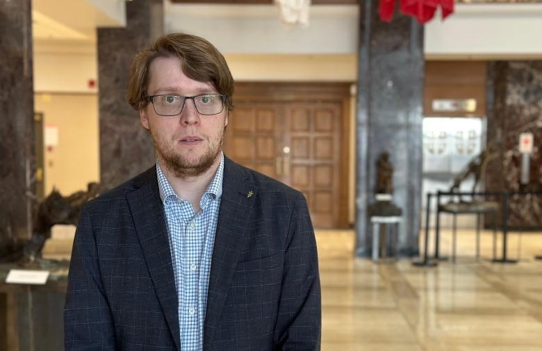 A man with brown hair, glasses, wearing a dark suit jacket and blue checkered shirt.