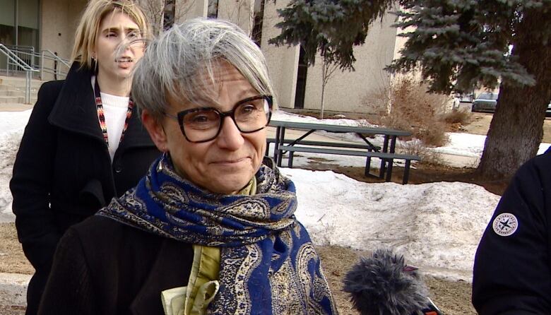 A woman with a scarf and glasses speaks into a microphone.