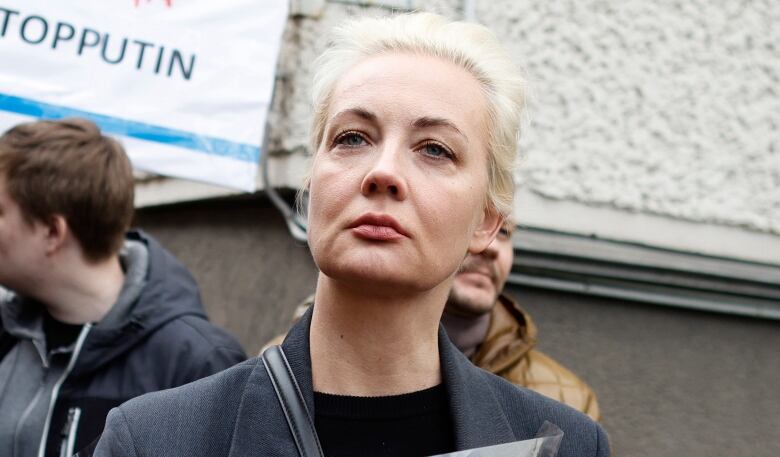 A blonde woman in a dark overcoat looks skyward, standing outdoors amid a crowd.