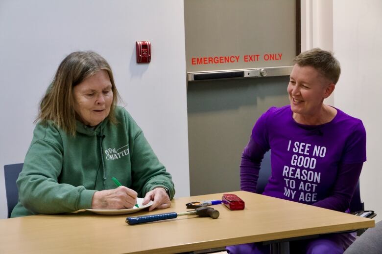 A senior woman write on a white plate with a permanent market while another woman wearing a T-shirt that says 'I see no good reason to act my age' stares on. A hammer sits on the table in front of them.