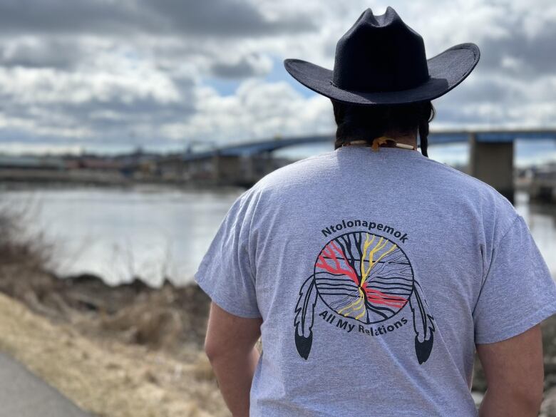 A man wearing a black cowboy hat and gray t-shirt stands outside with his back to the camera, looking out at a bridge over a river.