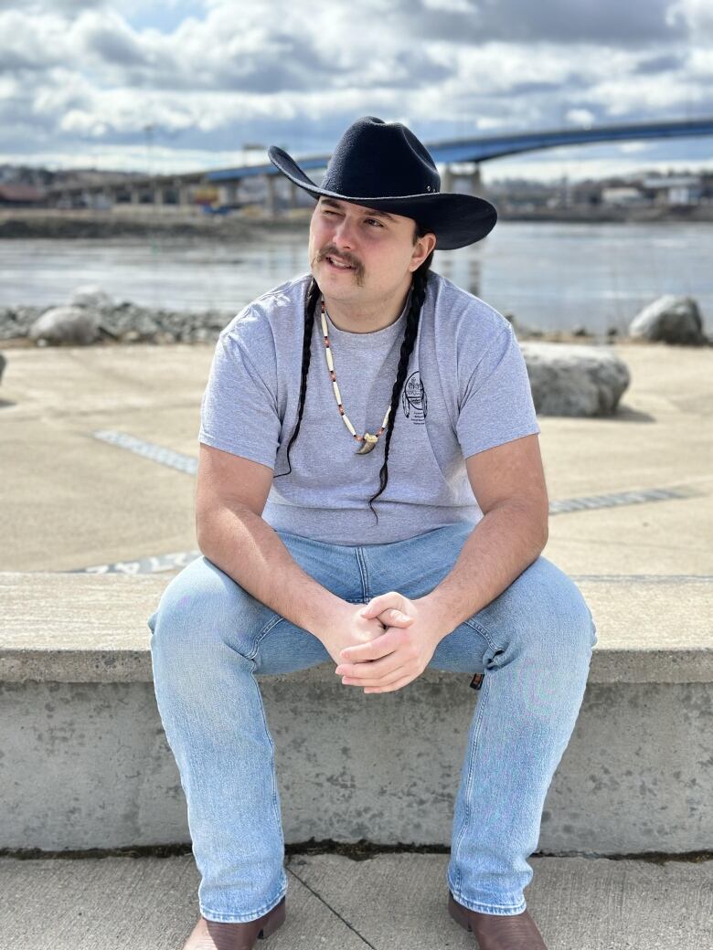 A man with a moustache and two long braids wearing a black cowboy hat sits on a cement bench. In the distance is a river and a bridge.