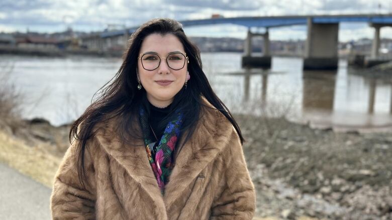 A young woman with long, dark hair wearing a brown fur coat stands outside in front of a river.