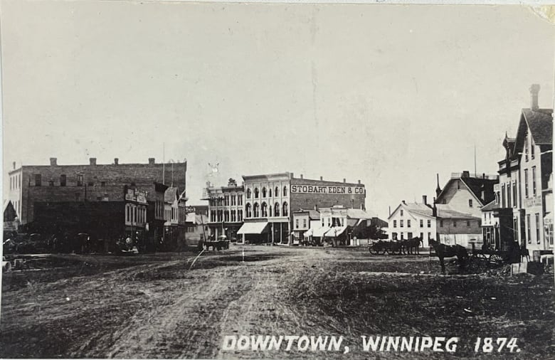 Black and white photo shows a wide muddy street, some horse carts and a few buildings, none more than three-storeys tall.