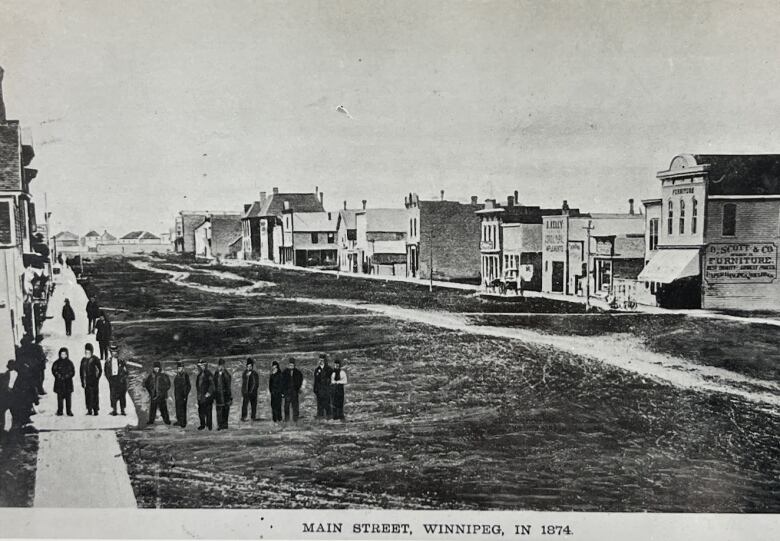 Black and white photo of a wide, muddy road bordered by a few wooden buildings. A crowd of about a dozen people are seen on a wooden plank sidewalk as well as the road.