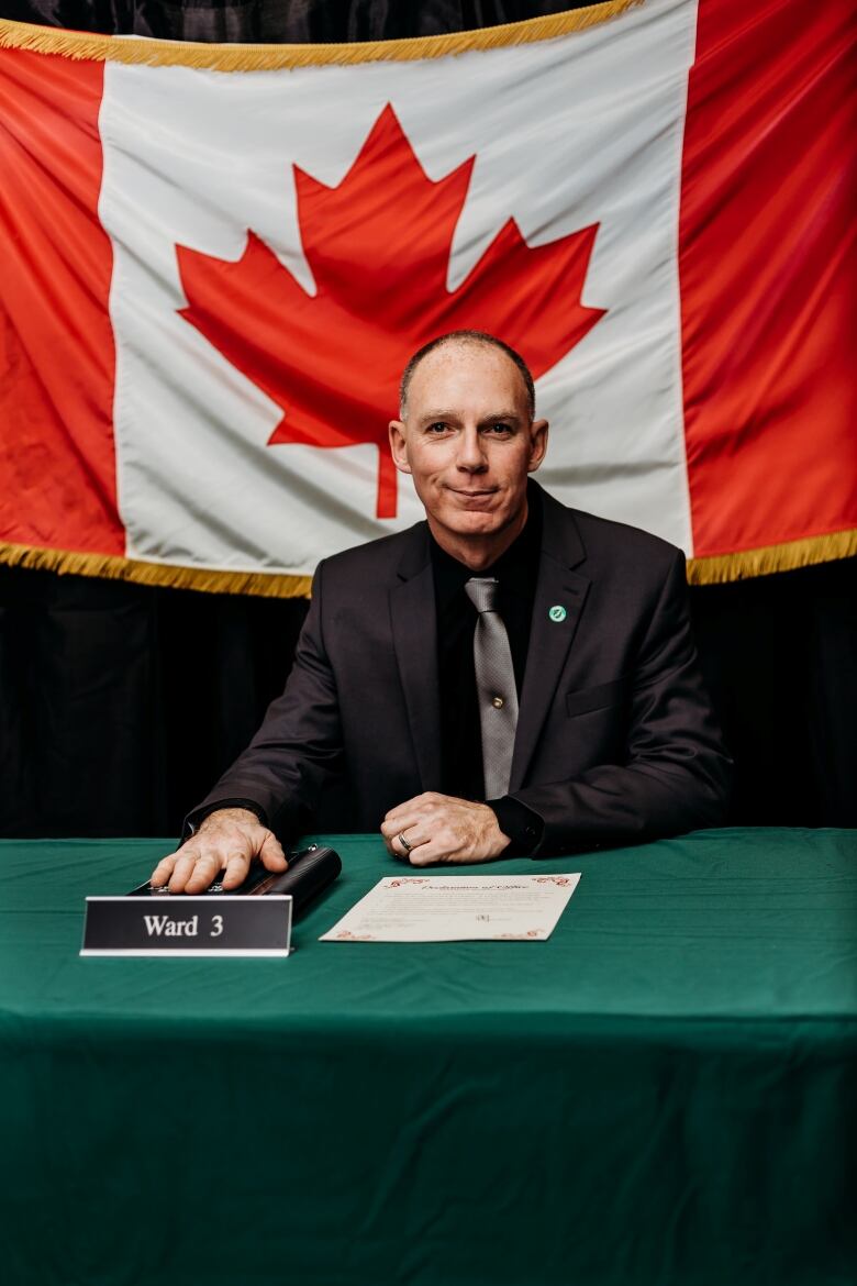 A man in black suit sits at a table with his hand on a bible, with a Canadian flag hung behind him.