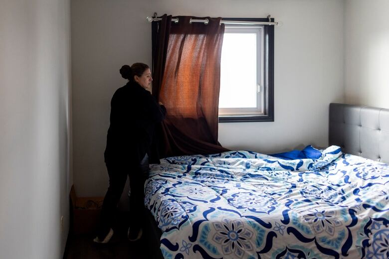 A woman adjusts the curtains in a sparsely furnished bedroom.