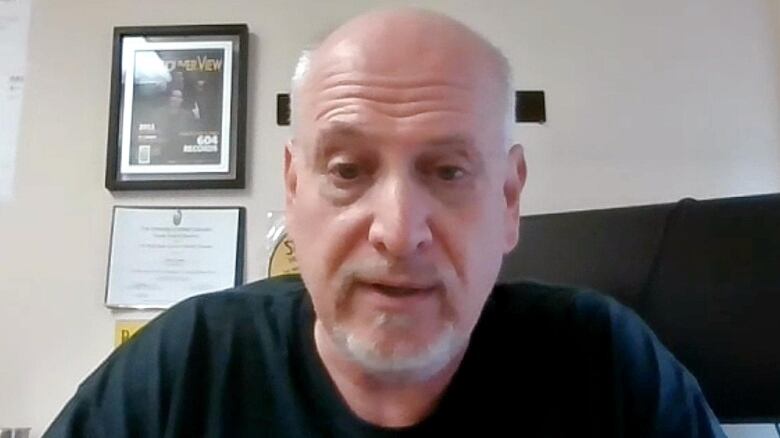 A man wearing a dark t-shirt is seated in front of a computer with framed pictures on a white wall behind him. 