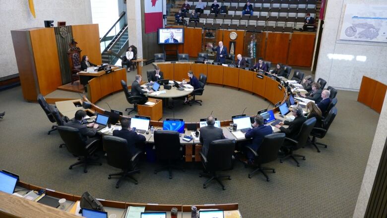 A large ornate room has desks arranged in a circle. A large canadian flag is draped on one wall. There are people sitting in a gallery in the back.