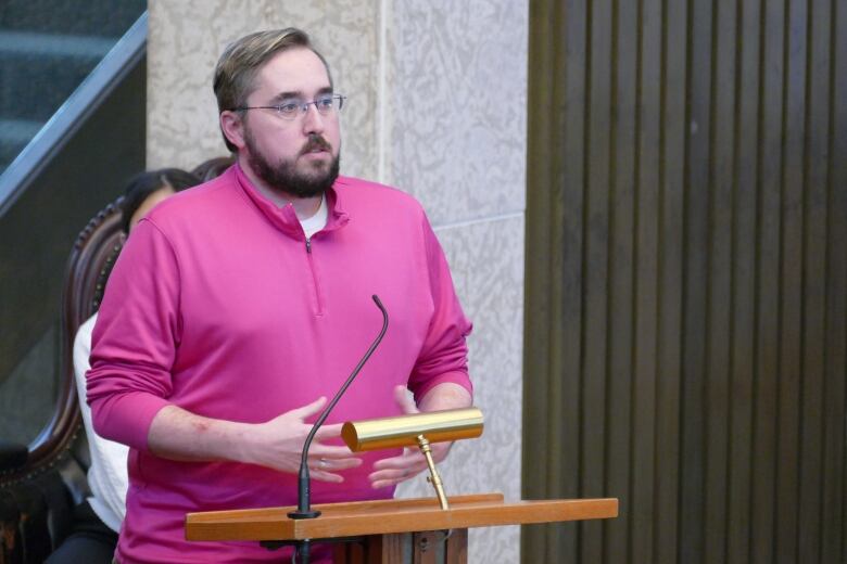 A man with a beard and wearing a pink shirt is standing and speaking at a podium.