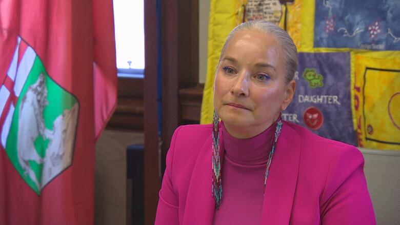 A woman in a pink suit and turtleneck is seated at a chair, in front of a quilt and a flag of Manitoba.