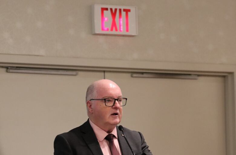 A man with black framed glasses wearing a suit and tie speaks into a microphone. Above his head is a red and white exit sign positioned over two doors.