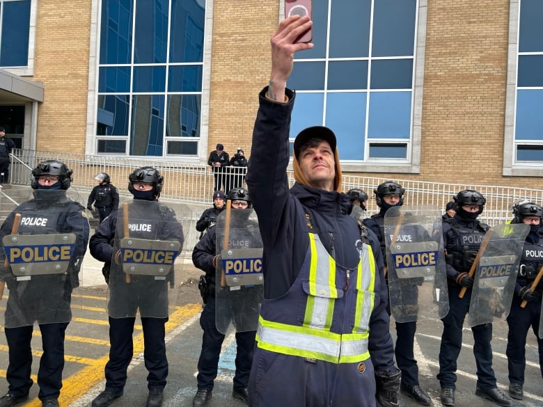 A man in work overalls holds his camera up, recording himself and a row of police officers in riot gear behind him.