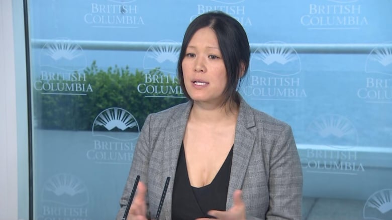 A woman speaks at a podium backed by British Columbia crests on a window.