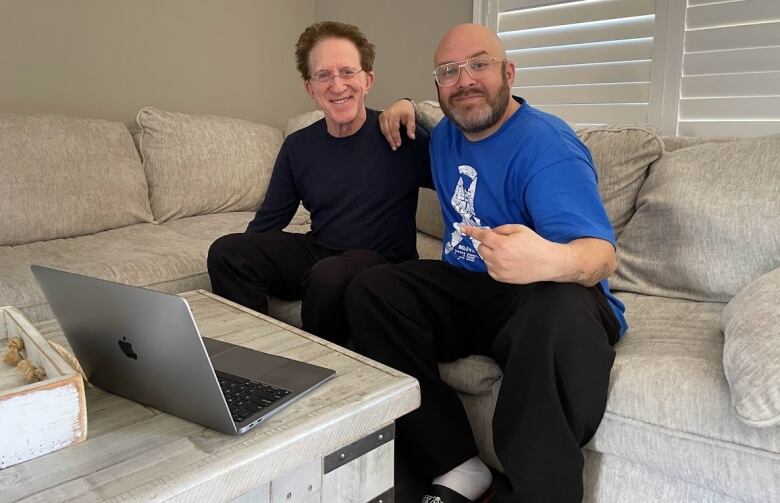 A man in a blue t-shirt sits with a man in a black long-sleeved shirt on a sectional sofa next to a coffee table with an Apple laptop.