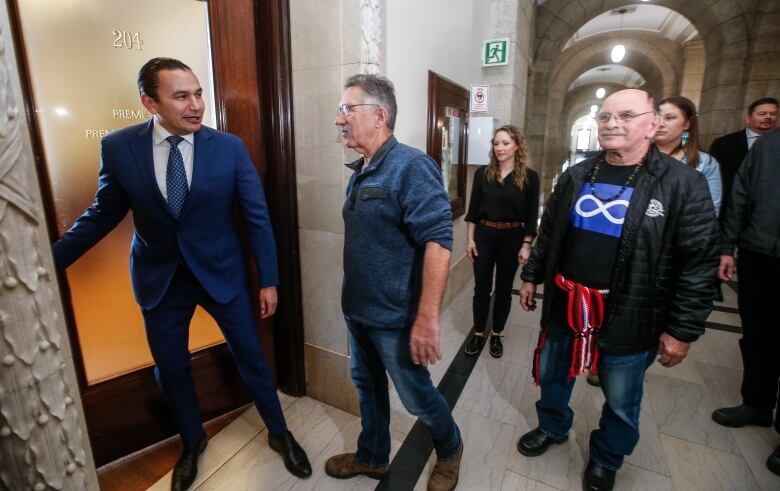 A man stands with his hand on a doorknob, two men next to him, with others behind him in a hallway.