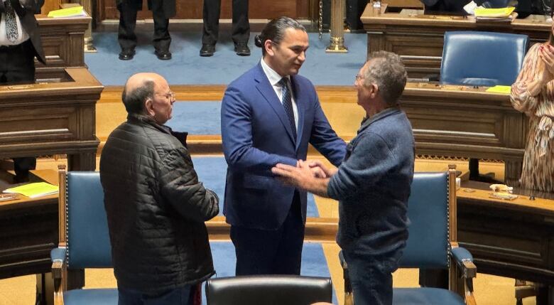 Two men stand facing a third man, who faces the camera. He shakes hands with the man on the right. On the edges of the photo are tiered desks, with people standing and applauding.