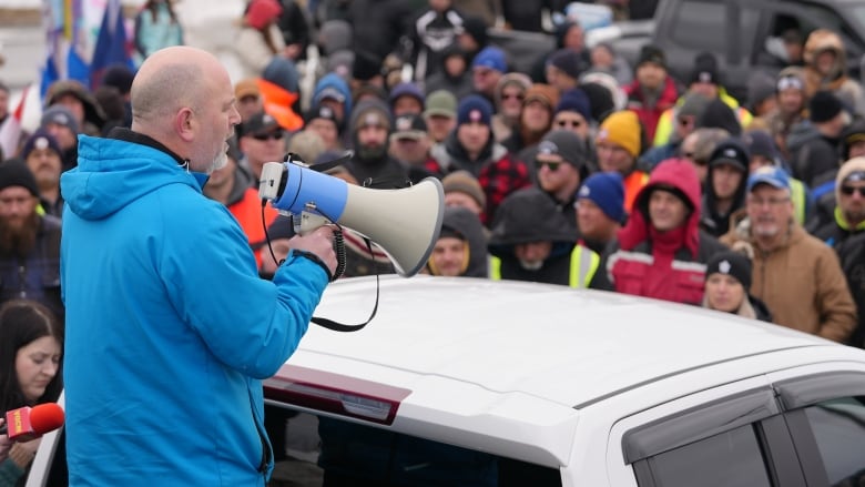 A man in a blue jacket holding a megaphone with a crowd in front of him.