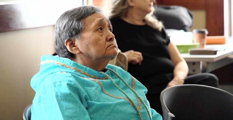 An elder sits in a chair wearing a traditional Inuit coat. 