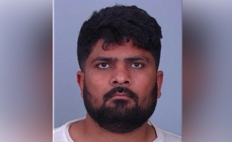 A mugshot of a South Indian man with a black beard wearing a white t-shirt, posing expressionless in front of a plain background.