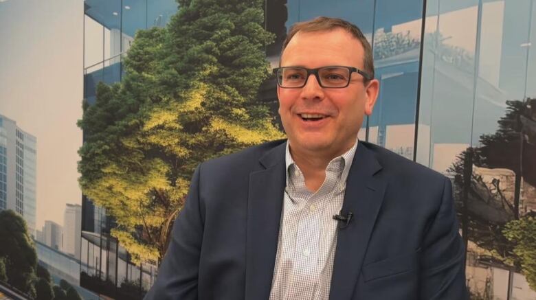 A man in a suit is interviewed in from of a banner showing office towers and trees.