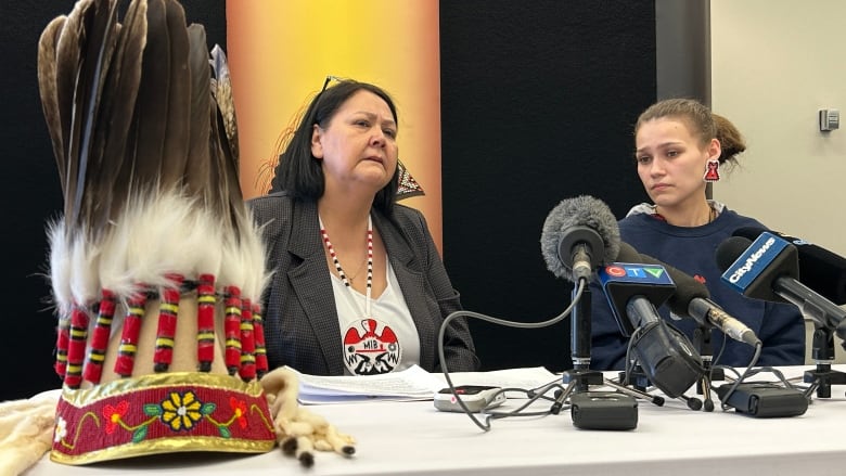 Two women sit behind a table with an array of microphones on it.