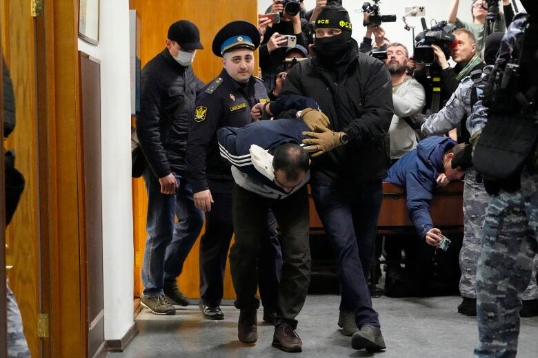 A person with their hands handcuffed behind their back is forcefully guided by police officers through a court house as photographer take pictures.