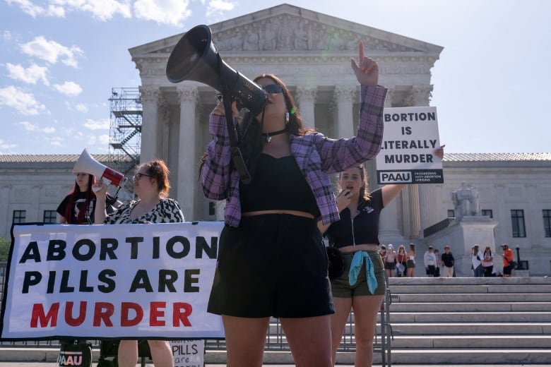 A woman wearing sunglasses, a flannel shirt and shorts is shown speaking into a megaphone at a demonstration in front of the steps leading to an august building. Near her, a sign reads, 'Abortion pills are murder.'