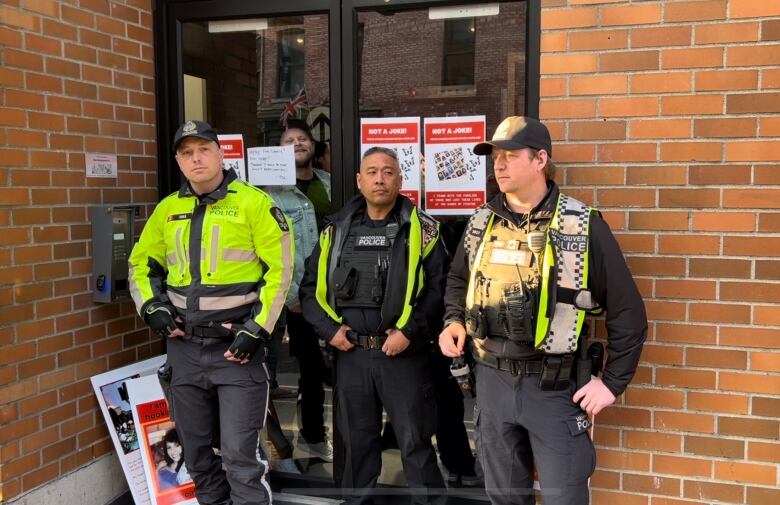 Three police officers wearing yellow vests stand in front of a door.