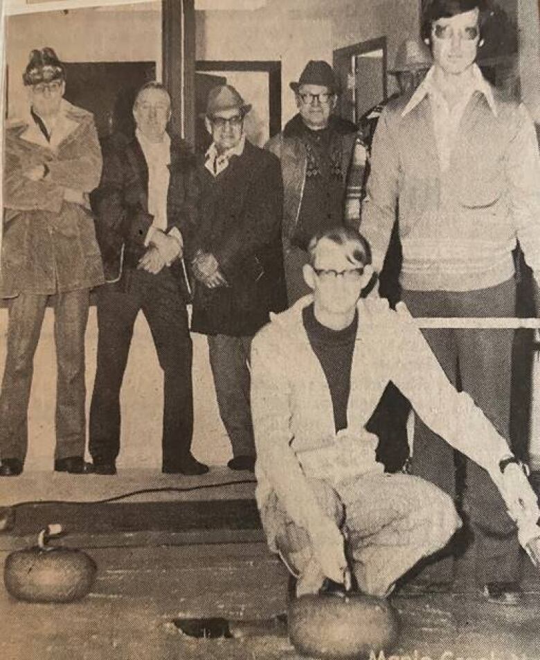 A grainy black and white photo shows a man hunched over a curling rock while five others watch nearby.