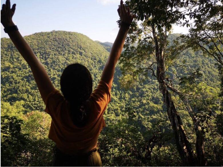 Woman, black hair, back to camera, orange shirt, with her arms held in the air looking into a lush green mountain range. 