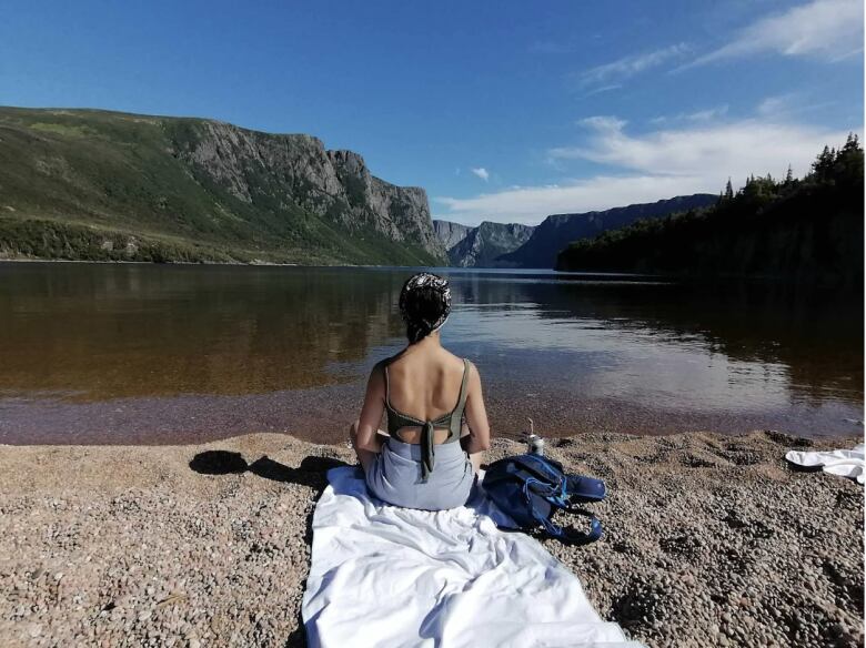 Woman sitting on a white blanket a beach, with back to camera. She's looking out to a mountain range and a blue sky with light clouds.