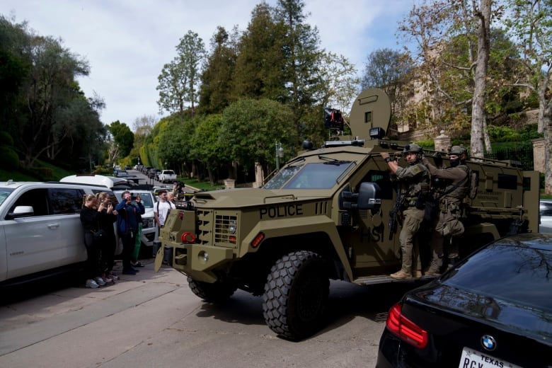 Two men in fatigues hold onto the side of a large military style vehicle as it drives around other stopped cars.