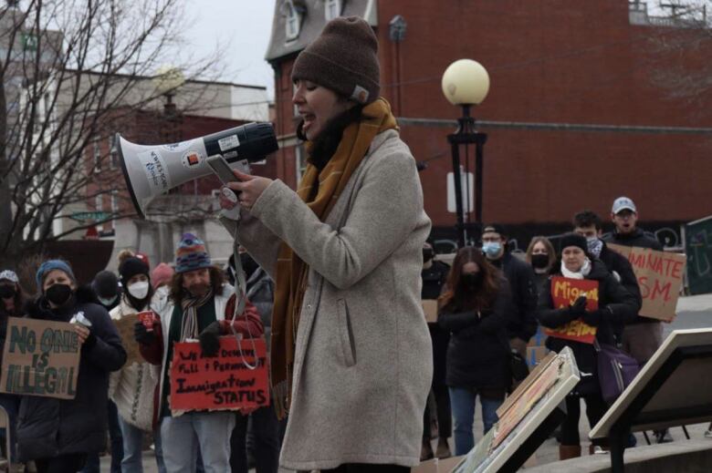 Woman in beige coat and brown hat speaks to people at a rally through a megaphone.