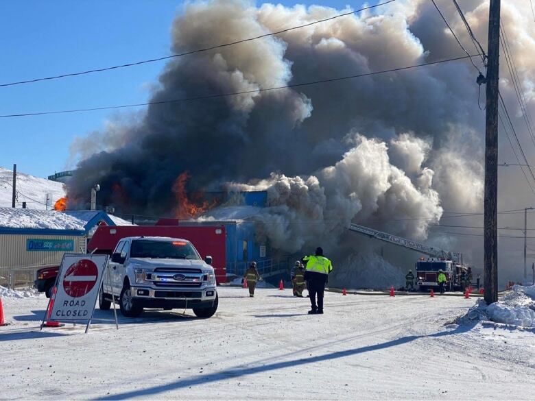 Black smoke and orange flames come out of a blue building with first responders in the foreground