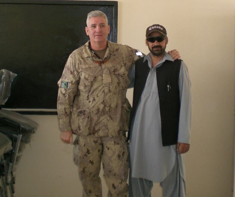 Two men one in a Candian military uniform, the other in traditional Afghan dress, stand side by side in a classroom.