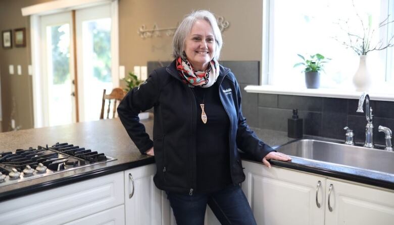 A woman with short light hair in a black vest and scarf stands in a bright kitchen near a window. 