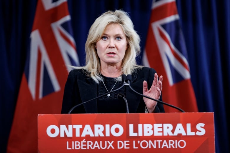 A woman stands behind a podium staring forward, her left hand is raised.